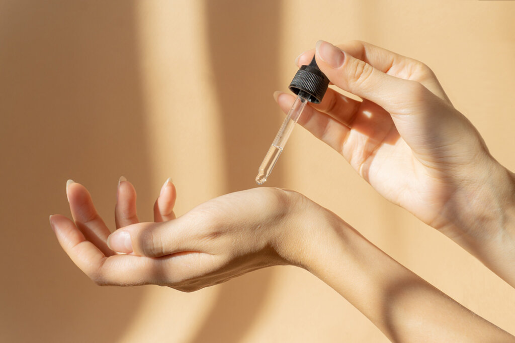 A woman’s hands are shown as she applies a skincare product to her palm using a pipette from a glass bottle.