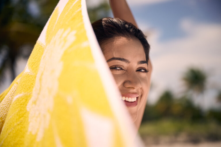 A smiling woman at the beach, partially covered by a yellow towel with a tropical background, enjoying a sunny day.