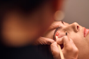A woman is getting her brows waxed. A Cerologist applies wax just below the brow line.