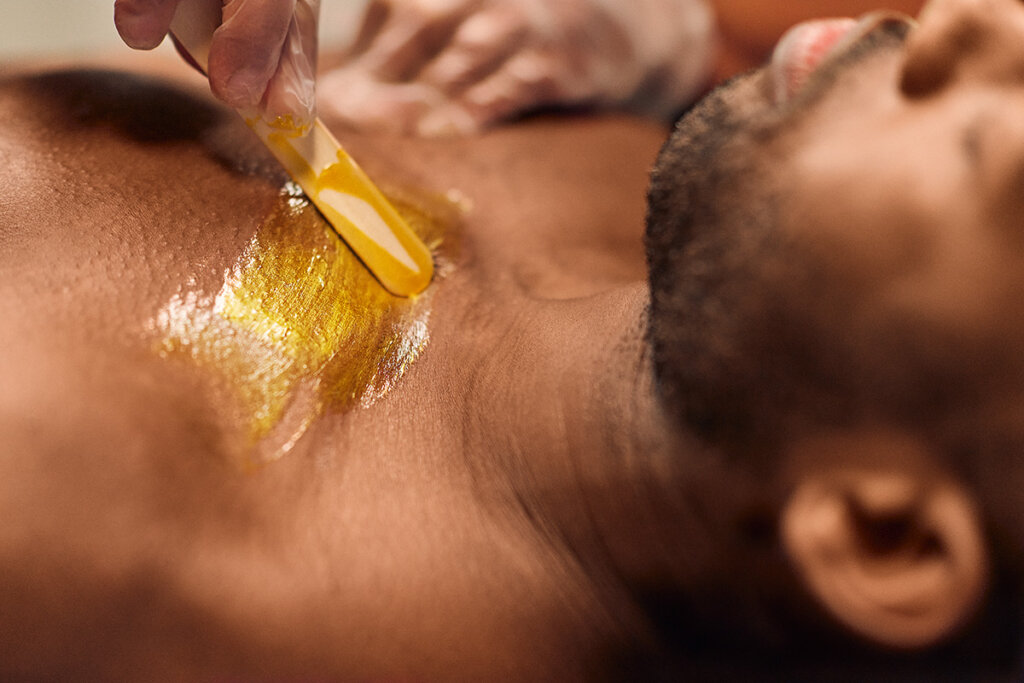 A man is having his chest waxed. A Cerologist applies wax to his skin using a wooden stick.