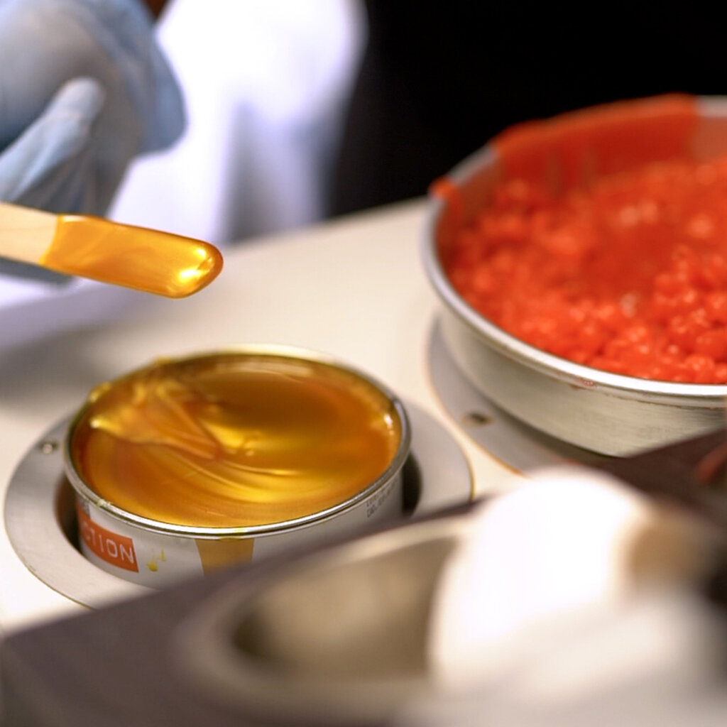 Hot orange wax is shown. Someone wearing gloves uses a wooden stick to stir and retrieve a small amount of wax.