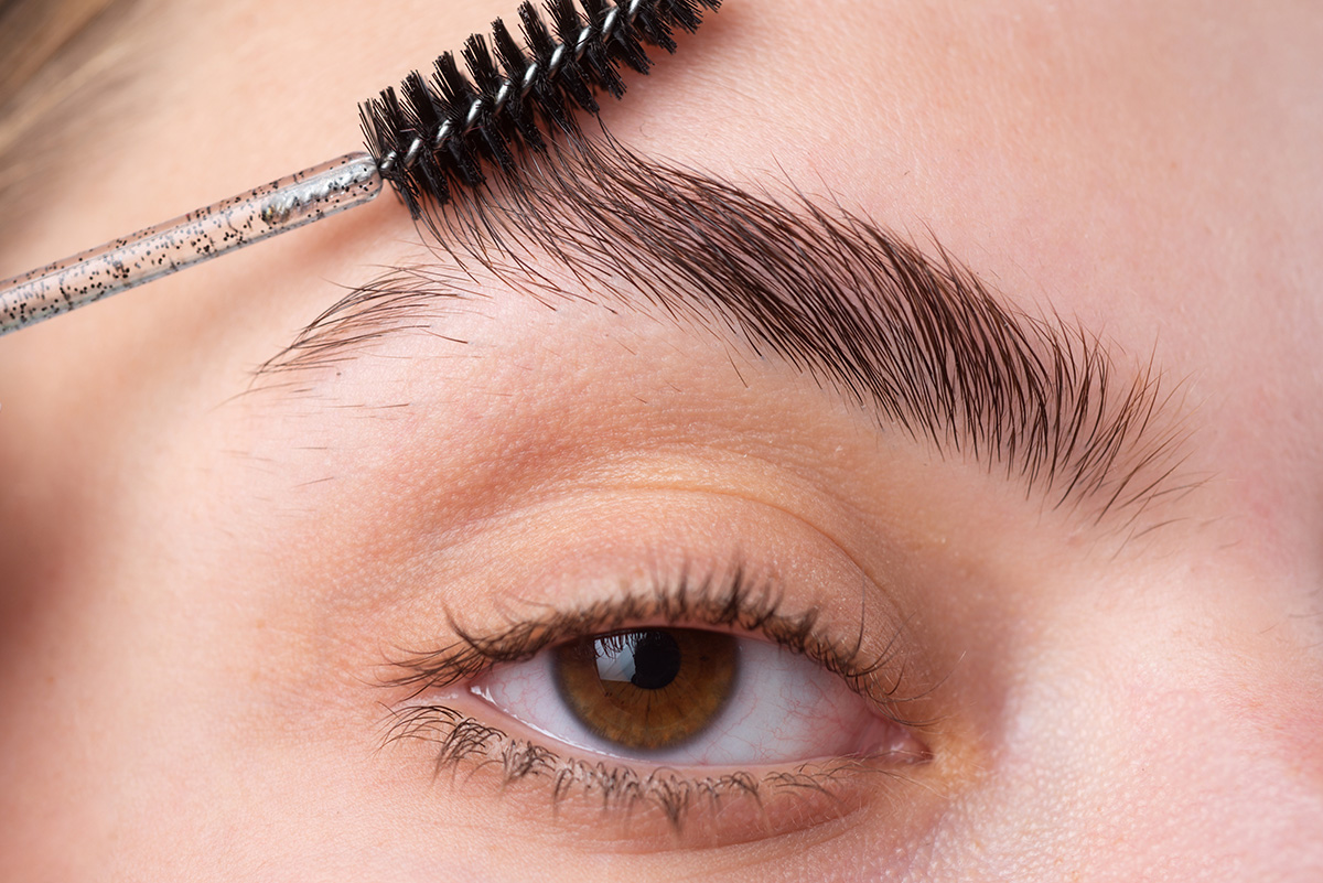 A woman’s eye and eyebrow are shown close up. Her eyebrow is being brushed with a small brow brush.