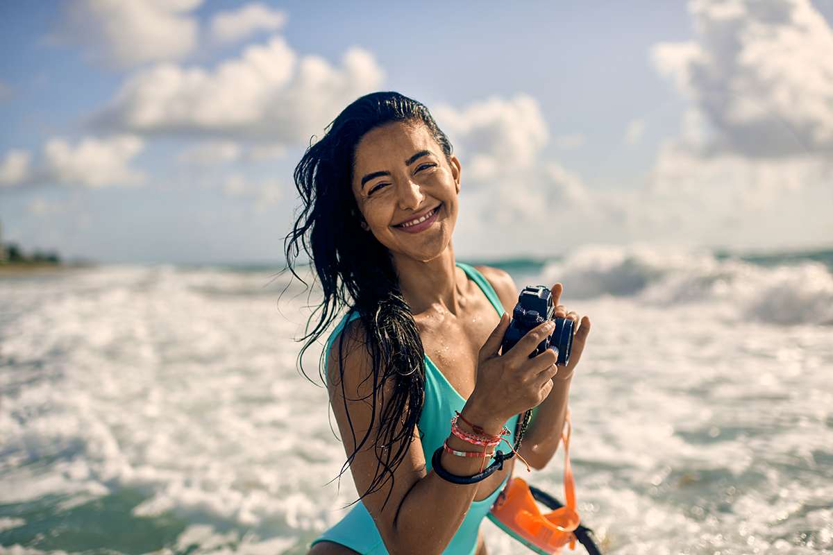 A woman is shown smiling in the water at the beach while holding a camera.
