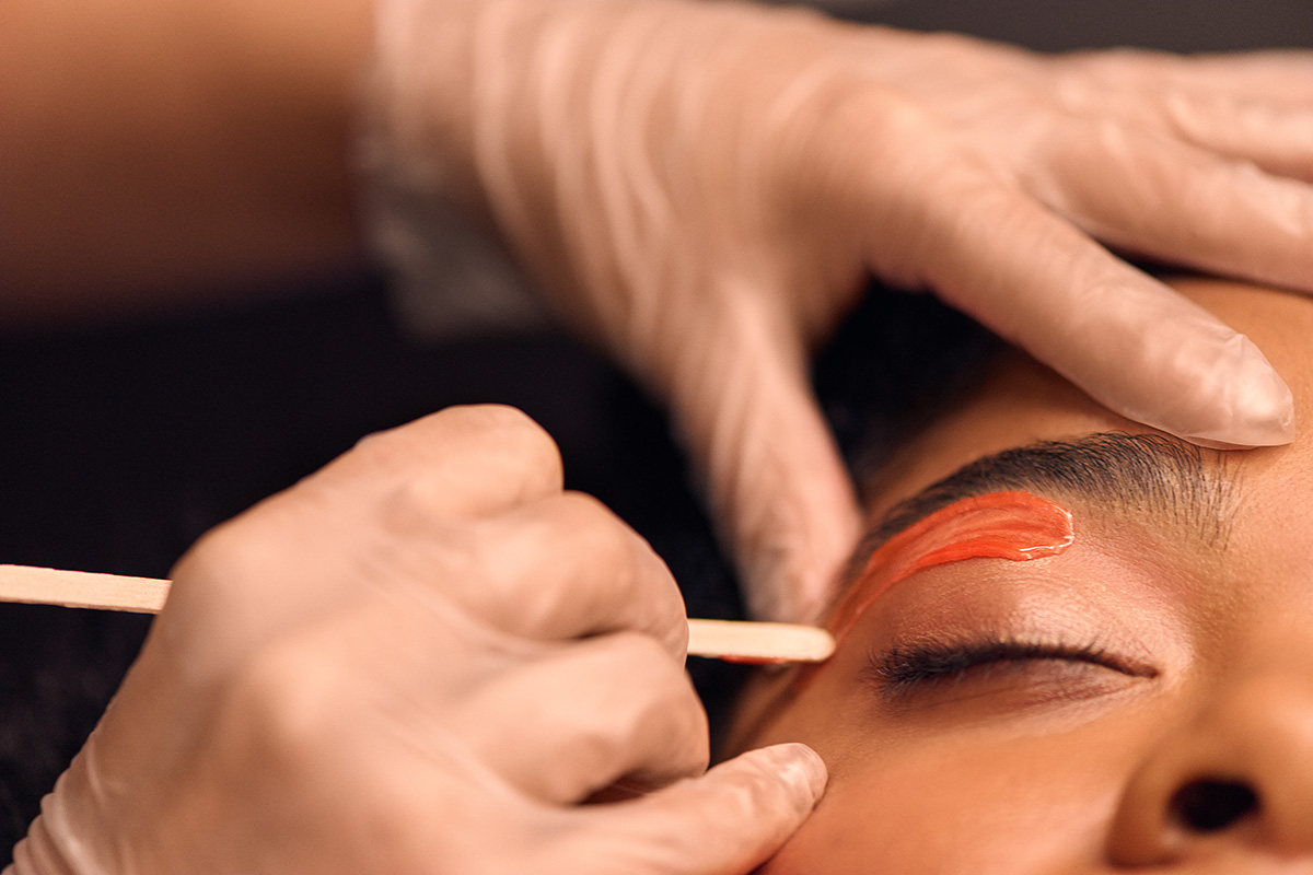 A woman is closing her eyes as her eyebrows are waxed by a professional who is wearing gloves.
