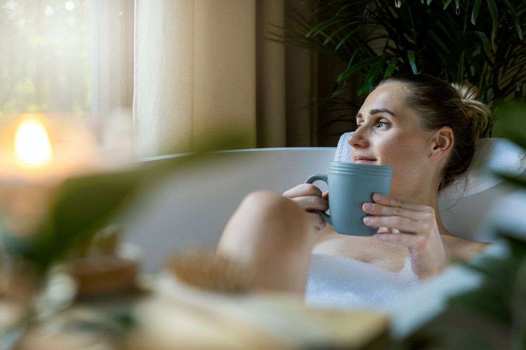 A woman is sitting in the bath tub while holding a mug and looking out the window.