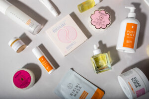 A flat lay of spring-themed self-care and skincare products from Waxing the City, including Post Wax Serum, Sugar Scrub, Bikini Mask, lip scrub, body oil, eye gels, and Spongellé body buffers on a light background.