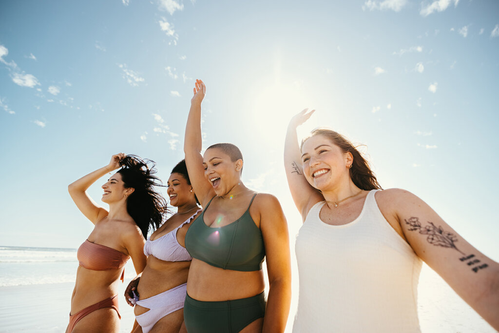 Four women in swimsuits walking and smiling near the ocean with the sun in the background.